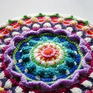 Puede incluir: Un mandala de ganchillo colorido con un arcoíris de colores, incluyendo rojo, naranja, amarillo, verde, azul, morado y blanco. El mandala tiene un diseño en capas con una flor central y múltiples filas de puntadas.