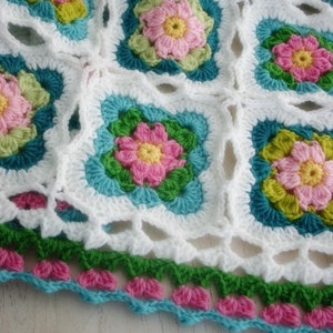Peut inclure: Une couverture au crochet avec un fond blanc et des motifs floraux colorés. La couverture a une bordure turquoise, rose et verte.