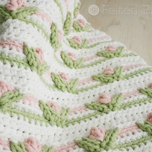 May include: A white crochet baby blanket with a repeating pattern of pink flowers and green leaves.