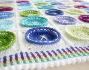 Könnte beinhalten: Eine weiße Häkeldecke mit einem bunten Knopfdesign. Die Decke ist in verschiedenen Farben gehalten, darunter Grün, Blau, Lila und Weiß. Die Knöpfe sind in die Decke gehäkelt und haben eine erhabene, strukturierte Oberfläche.
