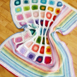Pode incluir: Uma manta de crochê colorida com uma borda de arco-íris. A manta é feita de quadrados com centros de diferentes cores e bordas brancas. A manta está dobrada ao meio e está sobre um piso de madeira.