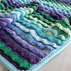 May include: A crocheted blanket with a wavy texture in shades of green, blue, and purple. The blanket has a light blue border and the text "Felted Button" is visible in the lower right corner.