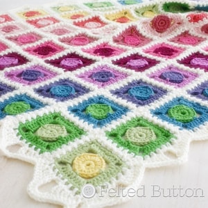 Op de afbeelding: Een regenboog gehaakte babydeken met een witte rand en een gegolfde zoom. De deken is gemaakt met een verscheidenheid aan kleuren, waaronder roze, paars, blauw, groen en geel. De deken is gemaakt met een oma-vierkant patroon en heeft een textuur. De deken ligt op een wit oppervlak. "Felted Button" staat in witte tekst op een witte cirkel.