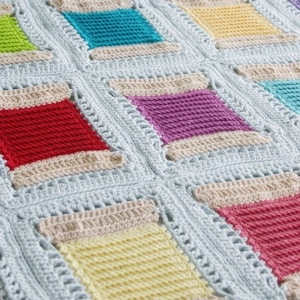 May include: A crocheted baby blanket with a light blue background and colorful squares. The squares are in shades of red, yellow, green, purple, and orange. The blanket is made with a simple stitch pattern and has a scalloped edge.