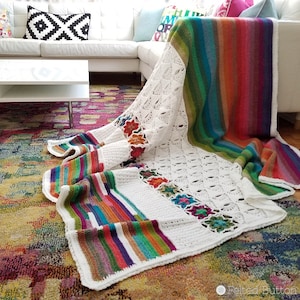 May include: A white crochet blanket with a colorful striped border. The blanket is draped over a white couch and a colorful rug.