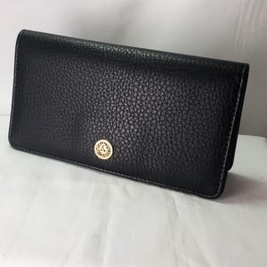 May include: Black leather wallet with a textured surface. The wallet has a gold-colored emblem on the front. The wallet is rectangular and appears to be closed.