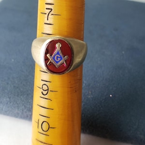 May include: A gold-toned ring with a red oval center featuring a Masonic symbol. The symbol includes a blue "G" within a square and compass design, with a ruler and other elements. The ring is displayed on a yellow measuring stick.