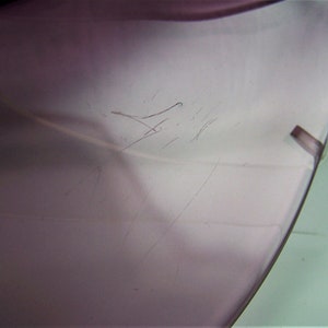 May include: Close-up of a light purple glass object with scratches on the surface. The object is slightly translucent and has a smooth, rounded edge.