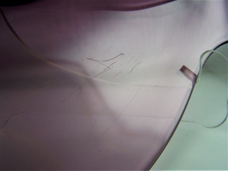 May include: Close-up of a light purple glass object with scratches on the surface. The object is slightly translucent and has a smooth, rounded edge.