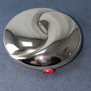 May include: A silver compact with a swirling design and a red jewel accent.