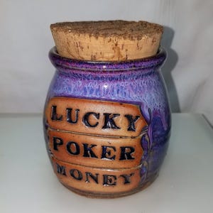 May include: A small, handcrafted ceramic jar with a cork stopper. The jar is glazed in shades of purple and blue, with the words "LUCKY POKER MONEY" in black lettering on a tan banner. A unique decorative item.