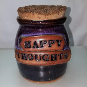 May include: A small, round, purple and brown ceramic jar with a cork lid. The jar has the words "HAPPY THOUGHTS" written on it.