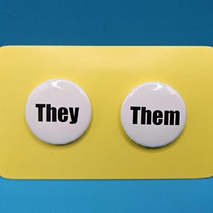 May include: Two circular white pin-back buttons featuring black text. The left button displays the word "They" and the right button displays the word "Them." Both buttons are mounted on a rectangular yellow card with rounded corners.