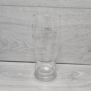以下が含まれることがあります： 幅広のベースとわずかに先細りのボディを備えた、透明で空のビールグラス。 グラスは透明なガラス製で、ビールを提供するように設計されています。 背景は明るい灰色の木製の表面です。