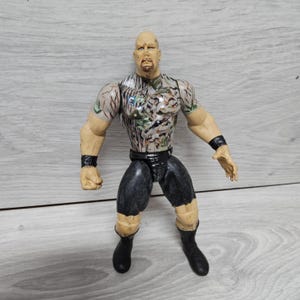May include: A plastic action figure of a bald man with a goatee, wearing a camouflage shirt, black shorts, and black boots. The figure has black wristbands and is posed with arms outstretched.