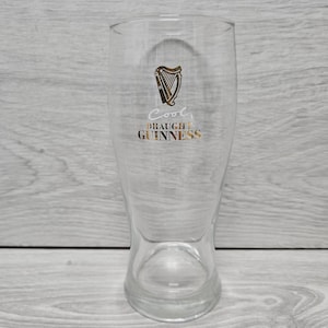 以下が含まれることがあります： 金色のハープロゴと「Cool Draught Guinness」の文字がプリントされた透明なパイントグラス。グラスは上部が広く、わずかにフレアしたベースがあります。グラスは空です。