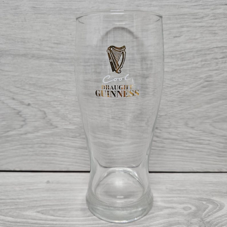 以下が含まれることがあります： 金色のハープロゴと「Cool Draught Guinness」の文字がプリントされた透明なパイントグラス。グラスは上部が広く、わずかにフレアしたベースがあります。グラスは空です。