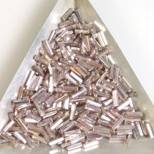 May include: A close-up of a pile of small, iridescent, light pink glass beads in a triangular metal tray. The beads are long and thin, resembling small tubes.