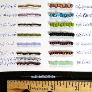 May include: A close-up of a white card with rows of different colored beads labeled with their size and type. The beads are arranged in rows, with the largest beads at the top and the smallest beads at the bottom. The card is labeled with the bead size and type, such as "15/0 Czech" and "6/0 Japanese".