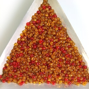 May include: A collection of small, round beads in shades of amber and red, filling a silver triangular tray. The beads are translucent, allowing light to pass through, and are likely used for jewelry making or crafting.