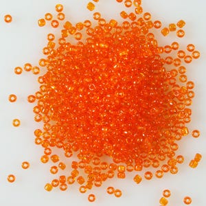 May include: A collection of small, translucent orange seed beads arranged in a loose, circular pile. Each individual bead features a smooth, rounded shape with a central hole, creating a vibrant and uniform display of bright orange colour.