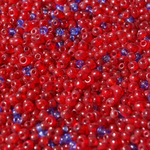 May include: A collection of small, round, translucent glass beads in vibrant red and deep blue colors. The beads are scattered together, creating a dense, textured surface with a glossy finish and visible holes in the center of each individual bead.