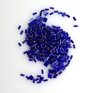 May include: A large pile of blue glass seed beads, scattered on a white surface. The beads are small and cylindrical, with a metallic sheen.