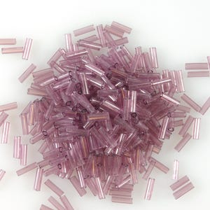 May include: A collection of translucent, pale purple glass tube beads. The beads are scattered on a white surface. The cylindrical beads are of varying lengths and are suitable for jewellery making or other craft projects.
