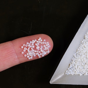May include: A close-up of a fingertip holding a pile of small, white, round beads. The beads are arranged in a loose pile, and some of them are overlapping. The beads are all the same size and shape, and they have a smooth, glossy finish.