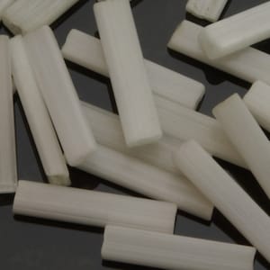 May include: A group of white glass beads, each shaped like a long, thin cylinder. The beads are scattered on a dark surface.