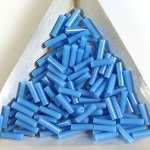 May include: A pile of light blue glass beads in the shape of cylinders. The beads are in a triangular metal tray.