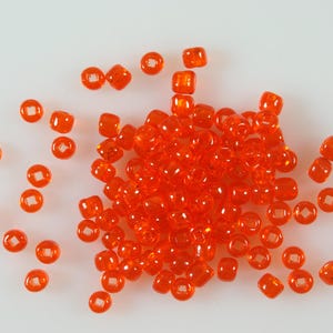 May include: A collection of small, translucent orange pony beads scattered across a surface. Each bead features a smooth, rounded shape with a clear, square-shaped hole in the centre, creating a vibrant and uniform appearance for crafting projects.
