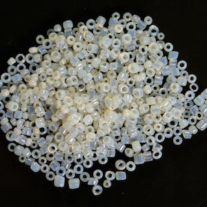 May include: A large pile of white and iridescent glass beads. The beads are round and have a hole in the center. They are scattered on a black surface.