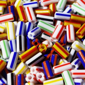 May include: A collection of cylindrical glass beads featuring a white base with vertical stripes in various colours including red, blue, yellow, green, and black. Each bead has a central hole running through its length, creating a multi-coloured pattern.