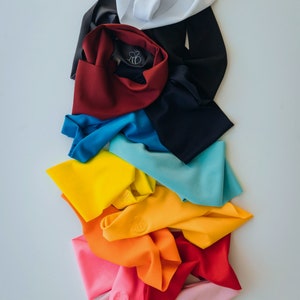 Op de afbeelding: Een stapel kleurrijke stoffen hoofdbanden met een bijenlogo. De hoofdbanden zijn gerangschikt in een regenboogpatroon, beginnend met wit en eindigend met roze.