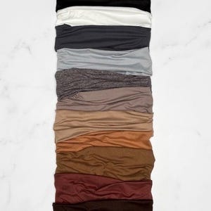 May include: A collection of twelve fabric headbands arranged in a vertical stack. The colors range from black, white, and gray to various shades of brown, tan, and deep red. Each headband features a smooth, flexible material with a soft texture.