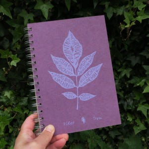 May include: A purple spiral bound notebook with a white leaf design. The leaf has words written on it in white, including "Elder" and "Trom".