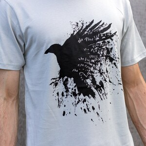 May include: A light gray t-shirt with a black crow graphic. The crow is surrounded by black paint splatters and the text "The Flos fit high" is repeated around the crow.