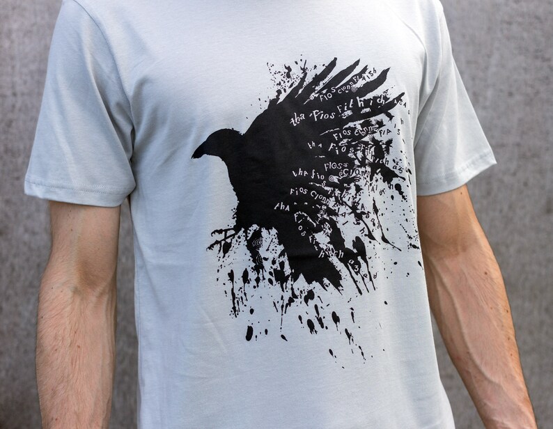 May include: A light gray t-shirt with a black crow graphic. The crow is surrounded by black paint splatters and the text "The Flos fit high" is repeated around the crow.