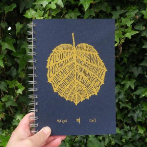 May include: A dark blue spiral bound notebook with a gold leaf design. The leaf is filled with Irish words. The words "Hazel" and "Coll" are printed at the bottom of the notebook.