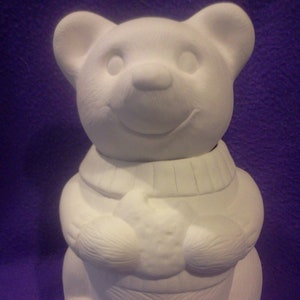 May include: White ceramic bear figurine with a smiling face, wearing a sweater and holding a star in its paws. The bear is sitting on a purple surface.
