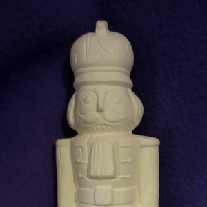 May include: White ceramic nutcracker figurine with a tall hat and a detailed face. The figurine is ready to be painted.