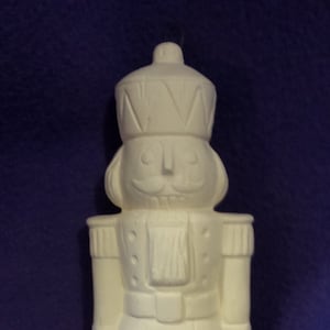 May include: A white ceramic nutcracker figurine, featuring a detailed design of a soldier with a crown, mustache, and uniform. The figurine is approximately 10 inches tall, perfect for holiday decor.