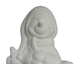 Może przedstawiać: Biała ceramiczna figurka bałwana z dzianym kapeluszem i szalikiem. Bałwan siedzi z założonymi nogami i ma uśmiechniętą twarz.
