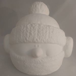 Op de afbeelding: Witte keramische figuur van een persoon met een gebreide muts en een sjaal. De figuur heeft een rond gezicht met een kleine neus en een brede glimlach.