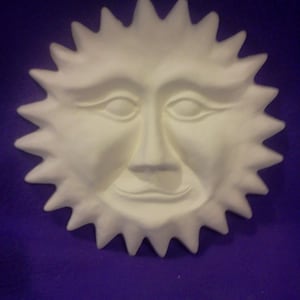 May include: White ceramic sun face wall decor with a smiling expression. The sun has a round face with a nose, eyes, and mouth. The sun has a spiky, starburst design around the edges.