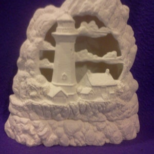 May include: White ceramic lighthouse figurine with a rocky base and clouds carved into the background. The lighthouse has a small house at its base and is surrounded by a rocky cliff.