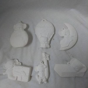 May include: Six white ceramic ornaments featuring a cartoon mouse design. The ornaments depict a mouse holding a present, a mouse in a hot air balloon, a mouse holding a candy cane, a mouse holding a book, a mouse in a crescent moon, and a mouse holding a bag.