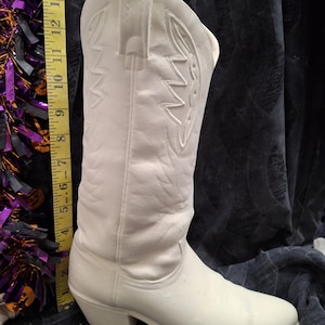 May include: A white ceramic cowboy boot figurine featuring embossed western-style stitching patterns on the shaft. A yellow measuring tape is positioned vertically next to the boot, indicating a height of approximately 14 inches from base to top.