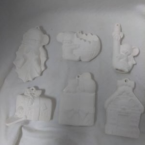 May include: Six white ceramic ornaments, ready to be painted. The ornaments include a house, a mailbox, a stocking, a reindeer, a penguin, and a snowman.